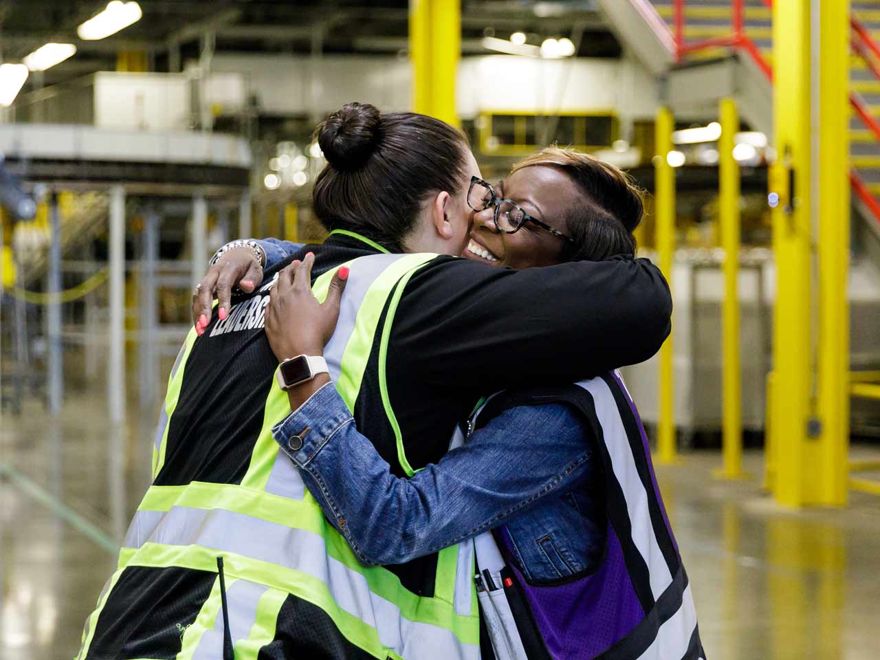 Two female Amazon employees wearing reflective vests share a warm hug standing on the production floor of an Amazon warehouse