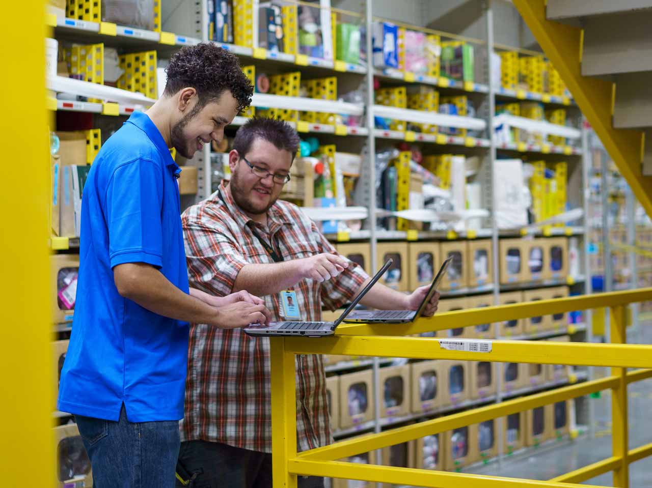 Two smiling Amazon employees stand with their laptops balanced on a safety railing on the production floor of an Amazon fulfillment center, with shelves of product in the background. One Amazon employee points to the other's laptop screen, teaching his colleague something.