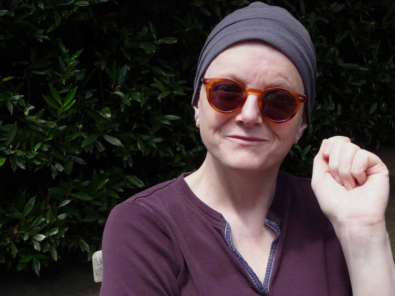 A woman wearing sunglasses and a chemo patient's skullcap in front of a hedgerow looking at the camera.